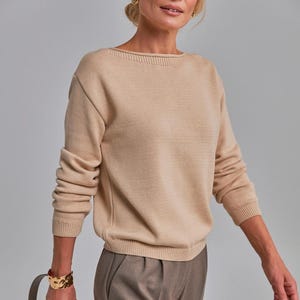 May include: A beige knit sweater with a boat neck and long sleeves. The sweater is paired with brown trousers. A gold bracelet and a tan handbag complete the look. The woman is wearing gold earrings.