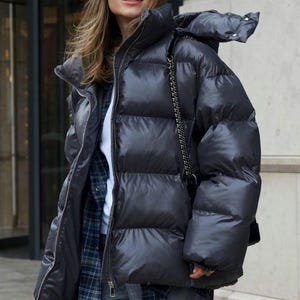 Könnte beinhalten: Eine anthrazitfarbene Daunenjacke mit hohem Kragen und Kapuze, getragen über einem weißen Hemd und einem blau karierten Hemd. Die Jacke hat einen Frontreißverschluss und eine schwarze Tasche mit Kettengurt wird über der Jacke getragen.