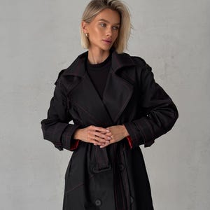 May include: A black trench coat with red stitching and a belt. The coat has a double-breasted closure and a classic collar.