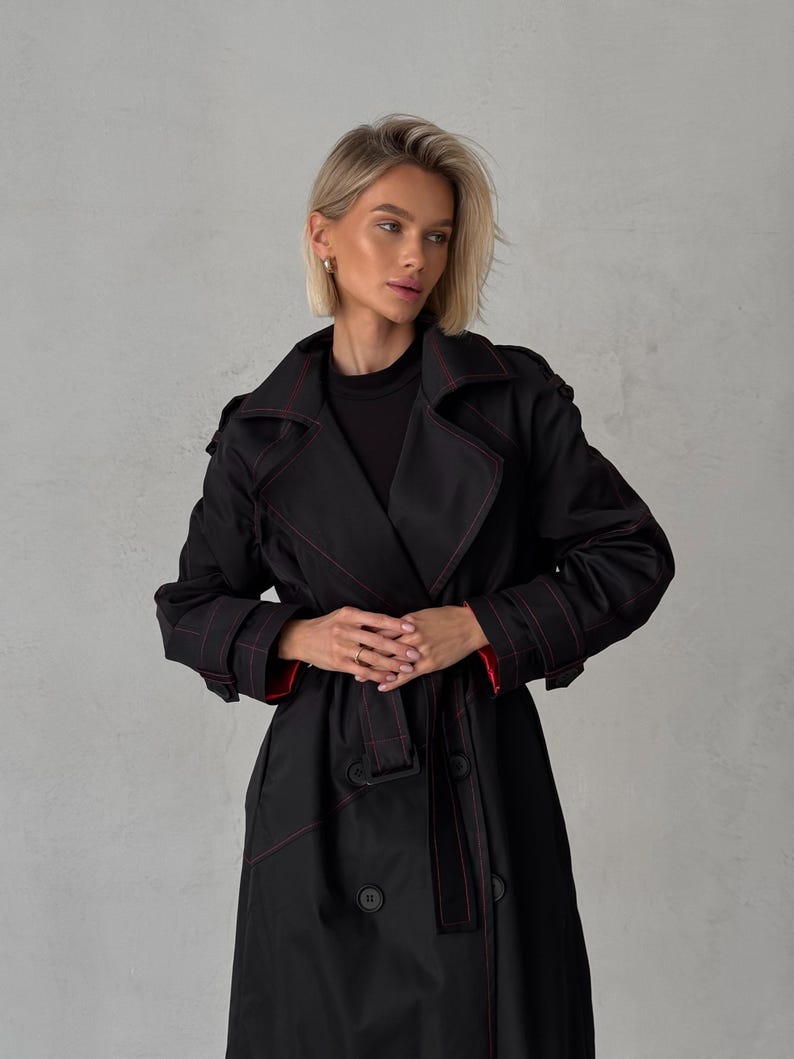 May include: A black trench coat with red stitching and a belt. The coat has a double-breasted closure and a classic collar.