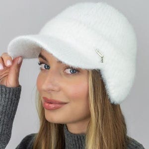 May include: A white, fluffy baseball cap with a curved peak. The cap has a soft, textured appearance and a small silver tag on the side. The cap is made of a fuzzy material, and is a winter accessory.