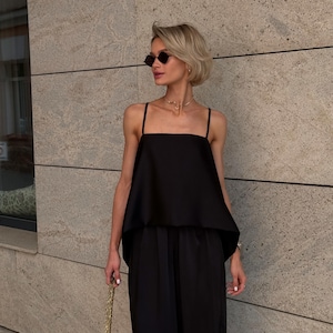 以下が含まれることがあります： 黒い服装の女性。四角いネックのトップスとワイドレッグパンツが含まれています。彼女はまた、黒いサングラスと金のネックレスを着用しています。金のチェーンが付いた黒いハンドバッグが彼女の手の中にあります。