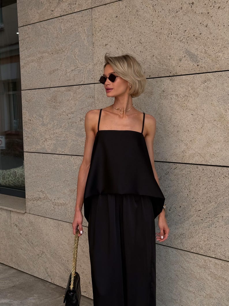 以下が含まれることがあります： 黒い服装の女性。四角いネックのトップスとワイドレッグパンツが含まれています。彼女はまた、黒いサングラスと金のネックレスを着用しています。金のチェーンが付いた黒いハンドバッグが彼女の手の中にあります。