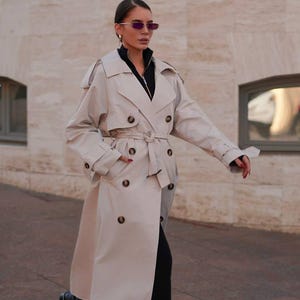 May include: A beige trench coat with a double-breasted design, a belt, and large buttons. The coat is worn over a black top and pants, paired with black boots. The overall look is stylish and modern.