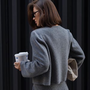 May include: A charcoal gray knit two-piece set consisting of a long-sleeved cropped sweater with a crew neckline and a matching mini skirt. The fabric has a soft, textured appearance, and the set is paired with a light beige clutch bag.