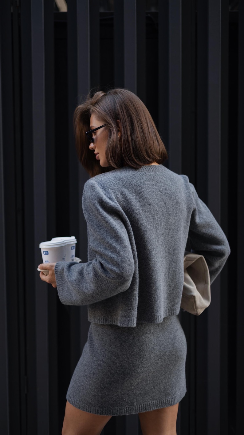 May include: A charcoal gray knit two-piece set consisting of a long-sleeved cropped sweater with a crew neckline and a matching mini skirt. The fabric has a soft, textured appearance, and the set is paired with a light beige clutch bag.