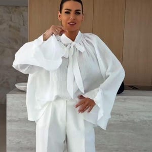 May include: A white blouse and pants set. The blouse features a high neckline with a tie, long sleeves, and a textured fabric. The pants are wide-legged. The outfit is all white.