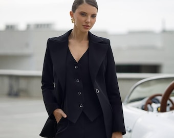 May include: A woman wearing a black suit with a vest. She is standing in front of a white car.