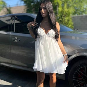Puede incluir: Un minivestido blanco con cuello halter y falda acampanada. El vestido se lleva con zuecos negros con plataforma. El fondo incluye un coche gris y un entorno soleado al aire libre.