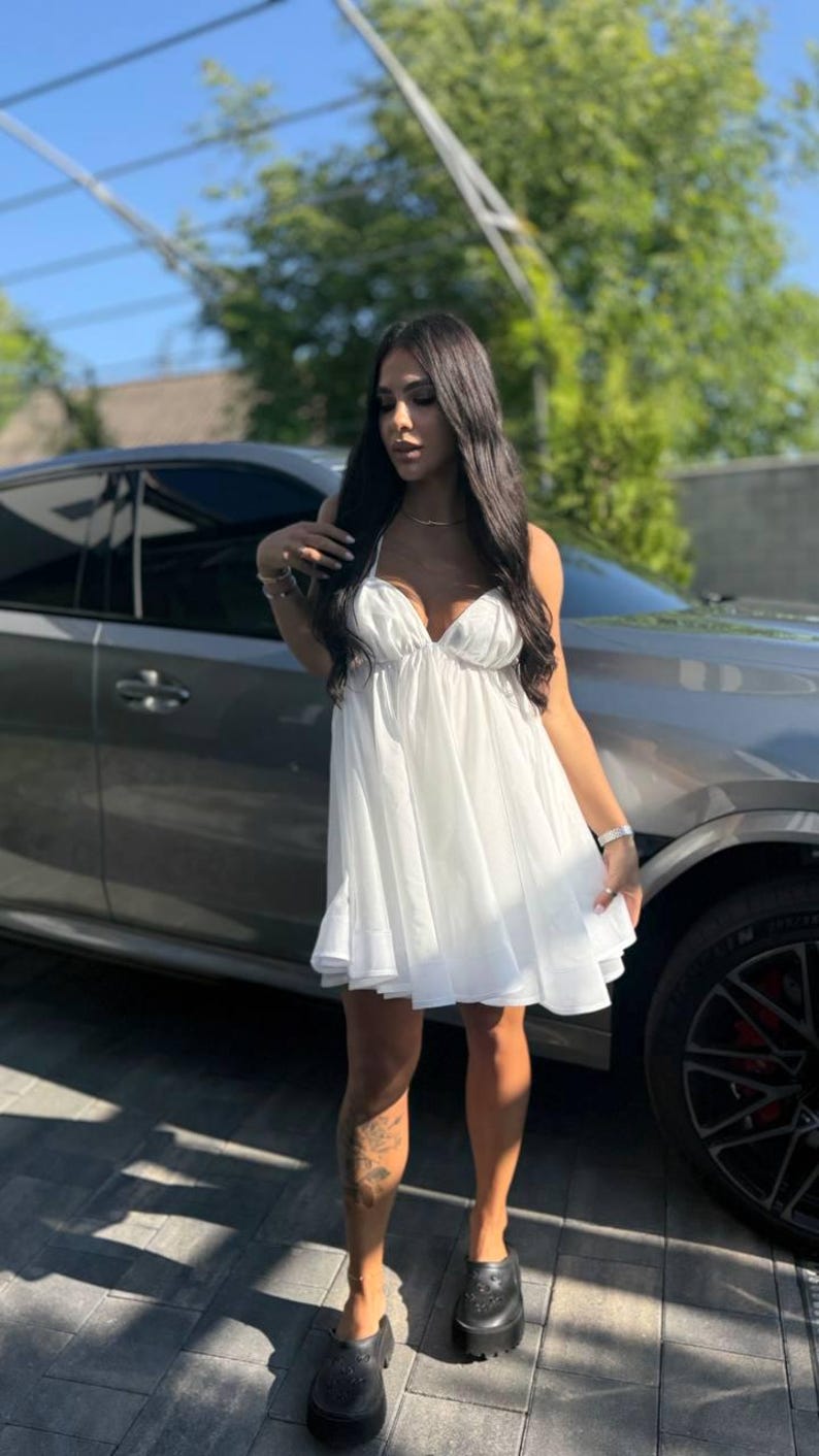 Puede incluir: Un minivestido blanco con cuello halter y falda acampanada. El vestido se lleva con zuecos negros con plataforma. El fondo incluye un coche gris y un entorno soleado al aire libre.