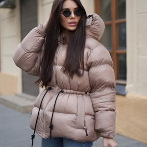 May include: A beige puffer jacket with a hood and a cinched waist, paired with blue jeans. The jacket has a drawstring detail and black drawstrings. The person is wearing black sunglasses and holding a brown purse.
