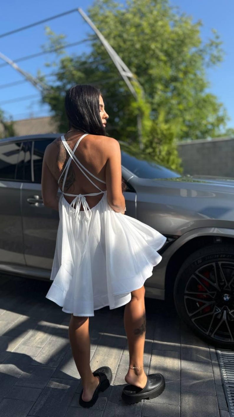 Puede incluir: Un vestido blanco con dise&ntilde;o de espalda cruzada. El vestido tiene una falda fluida y se sujeta ligeramente. La persona lleva sandalias de plataforma negras. Un coche gris est&aacute; en el fondo.