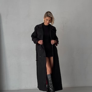 May include: A black long trench coat with a double breasted closure and a red lining. The coat is worn by a person with blonde hair and is paired with black knee-high boots.