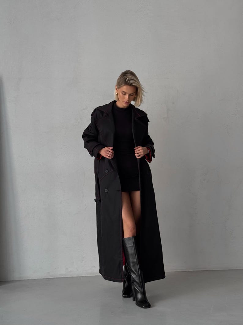 May include: A black long trench coat with a double breasted closure and a red lining. The coat is worn by a person with blonde hair and is paired with black knee-high boots.