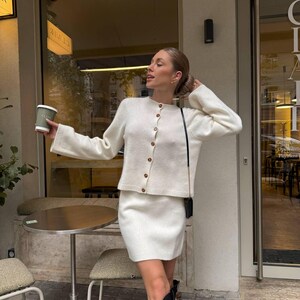 May include: A cream-colored knit cardigan with a round neckline and brown buttons, paired with a matching cream-colored knit mini skirt. The cardigan features long sleeves with ribbed cuffs, and the skirt has a simple, straight-cut hemline.