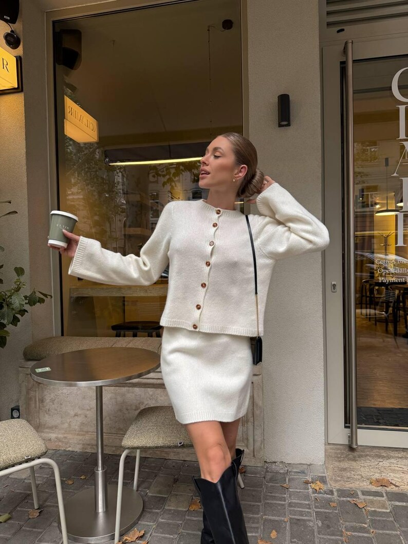 May include: A cream-colored knit cardigan with a round neckline and brown buttons, paired with a matching cream-colored knit mini skirt. The cardigan features long sleeves with ribbed cuffs, and the skirt has a simple, straight-cut hemline.