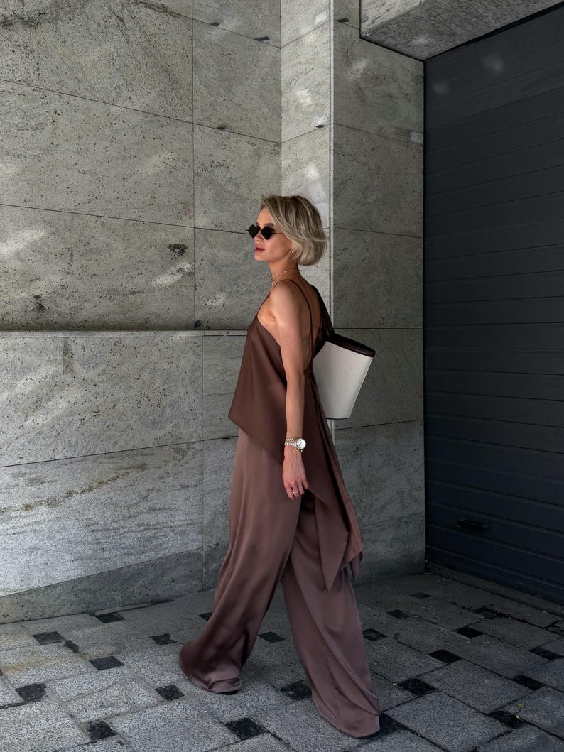 以下が含まれることがあります： 茶色のシルクのトップスと、お揃いのワイドパンツを着用した女性。黒いストラップの白いトートバッグ、黒いサングラス、シルバーの腕時計を身につけています。この服装はモノクロです。