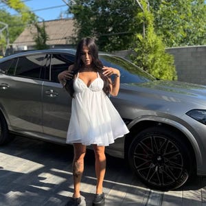 Puede incluir: Un vestido blanco, corto y fluido con tirantes finos. El vestido tiene un escote en V y una falda acampanada. La persona lleva zapatos negros. Un coche gris est&aacute; aparcado detr&aacute;s de la persona.