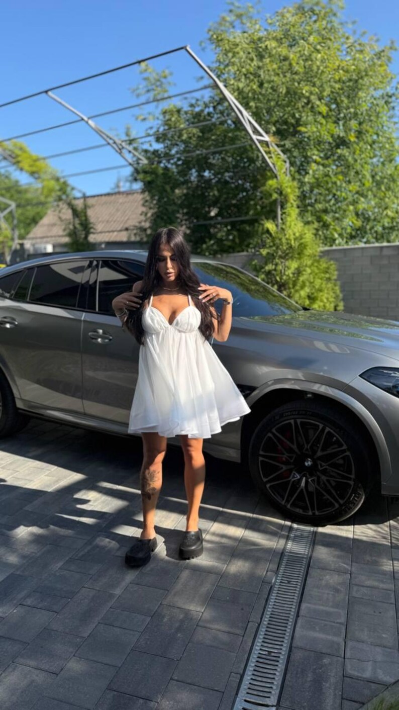 Puede incluir: Un vestido blanco, corto y fluido con tirantes finos. El vestido tiene un escote en V y una falda acampanada. La persona lleva zapatos negros. Un coche gris est&aacute; aparcado detr&aacute;s de la persona.