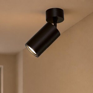 May include: A matte black cylindrical ceiling spotlight fixture with a swivel joint, mounted to a circular base. The light is angled downward, emitting a bright white glow from the open end of the cylinder, illuminating the space below.