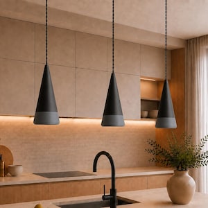 Può includere: Tre lampade a sospensione a forma di cono con sezioni superiori nero opaco e fasce inferiori grigie pendono sopra un'isola cucina. L'isola presenta un piano di lavoro chiaro, un rubinetto nero, una ciotola di pere e un vaso con fogliame verde.