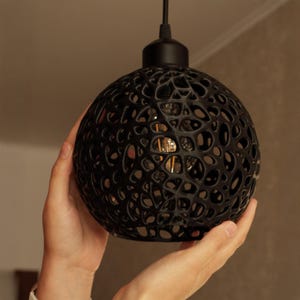 May include: A black, spherical pendant light fixture with a unique, open-weave design. The light fixture is held by two hands. The light bulb is visible through the openings. The fixture hangs from a black cord.