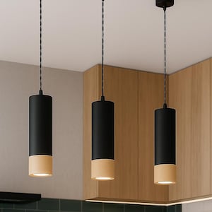 May include: Three modern pendant lights with a black cylindrical top and a beige bottom. The lights hang from the ceiling with black cords. The background features a kitchen setting with wooden cabinets and a green tiled wall.