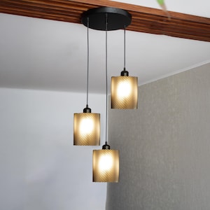 Puede incluir: Una moderna lámpara colgante de tres luces con pantallas cuadradas de vidrio texturizado. Las pantallas son de color ámbar y emiten un brillo cálido. La lámpara tiene una placa de techo negra y cables negros, creando una solución de iluminación elegante.