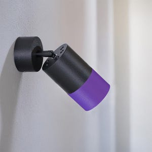Puede incluir: Un foco negro y morado montado en una pared blanca. La luz cilíndrica tiene un cuerpo negro con un extremo morado. El accesorio está diseñado para ser ajustable.