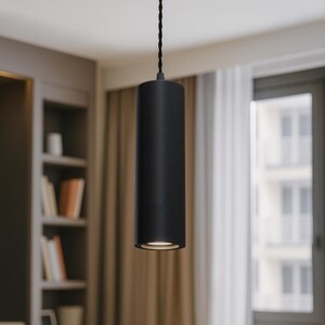 May include: A matte black cylindrical pendant light fixture suspended by a dark twisted fabric cord. The fixture features a clean, vertical tubular design with a circular opening at the bottom that emits a soft, focused downward light beam.