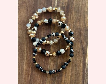 May include: Three beaded bracelets in a stack. The bracelets feature a mix of black, white, gold, and tan beads. Some beads are faceted, while others are smooth. The bracelets are arranged on a wooden surface.
