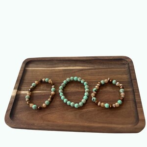 May include: Three beaded bracelets with turquoise and brown beads are displayed on a wooden tray. The bracelets have a mix of round and faceted beads, creating a textured and colorful accessory.