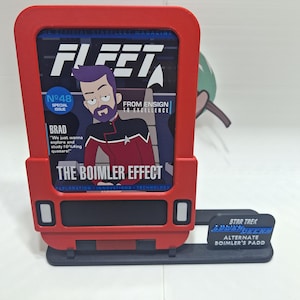 May include: A red and black Star Trek themed stand with a magazine cover featuring a cartoon character. The magazine cover reads "Fleet" and "The Boimler Effect". The stand also has a blue and white plaque that reads "Star Trek" and "Alternate Boimler's Padd".