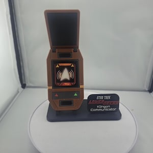 May include: A brown and black Star Trek Klingon Communicator prop. The prop has a flip-up screen and a display showing a white arrow and signal waves. The communicator sits on a black base with a Star Trek logo.