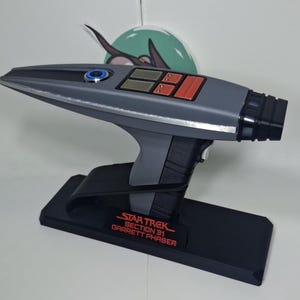 May include: A gray and black Star Trek Section 31 Garrett Phaser replica with a red button and a black stand. The stand has the words "Star Trek Section 31 Garrett Phaser" printed on it in red.