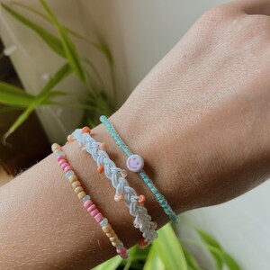 May include: Four colorful beaded bracelets. One bracelet is a light blue with a smiley face bead. Another is a braided white bracelet with orange beads. The third bracelet is pink and tan. The fourth bracelet is light blue.