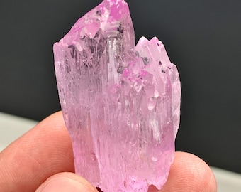 Könnte beinhalten: Ein roher, natürlicher rosa Kristallcluster mit einer kristallinen Struktur. Der Kristall hat ein durchscheinendes Aussehen mit verschiedenen Rosatönen. Der Kristall wird in einer Hand vor einem dunklen Hintergrund gehalten.