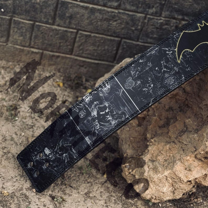 Peut inclure: Ceinture en cuir noir avec un design de bande dessin&eacute;e pr&eacute;sentant des illustrations de Batman. La ceinture a un logo Batman dor&eacute; et une boucle avec deux rivets m&eacute;talliques. La ceinture est pos&eacute;e sur un rocher.