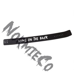 Peut inclure: Un bracelet noir avec le texte "NAME ON THE BACK" en blanc. Le bracelet comporte plusieurs trous pour le r&eacute;glage et deux trous &agrave; l'extr&eacute;mit&eacute; oppos&eacute;e. Le bracelet est fait d'un mat&eacute;riau souple.