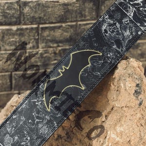 Peut inclure: Sangle de guitare noire avec le logo Batman bord&eacute; de jaune. La sangle pr&eacute;sente un motif de bande dessin&eacute;e en noir et blanc. La sangle est inclin&eacute;e sur une surface rugueuse et textur&eacute;e.