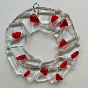May include: A circular wreath ornament constructed from clear, fused glass shards arranged in a mosaic pattern. The piece is accented with small, vibrant red glass fragments embedded throughout the clear structure and includes a metal hanging loop.