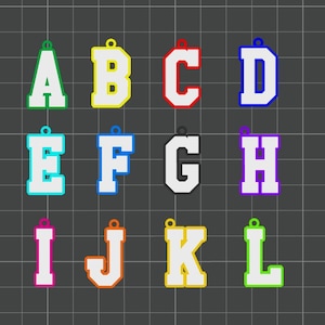 Peut inclure: Une collection de douze pendentifs en forme de lettres, chacun dans une combinaison de couleurs différente. Les lettres A à L sont affichées, chaque lettre ayant un centre blanc et un contour coloré. Les pendentifs sont disposés sur un fond de grille gris foncé.