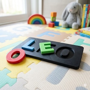 May include: A black wooden puzzle spelling out 'LEO' in blue, green, and red letters. The puzzle sits on a colourful foam play mat, with a rainbow, blocks, a stuffed bunny, and books in the background.
