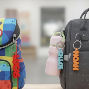 May include: A colorful backpack and a gray diaper bag are shown with personalized keychains. The keychains spell out the names "LUCAS", "CHLOE", and "NOAH" in red, blue, and orange. A pink water bottle is attached to the diaper bag.