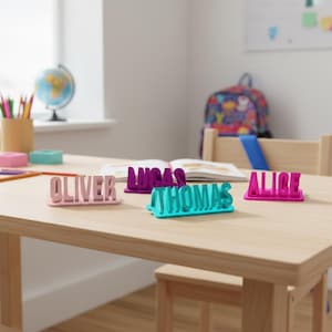 May include: A selection of brightly coloured 3D-printed nameplates spelling "OLIVER", "LUCAS", "THOMAS", and "ALICE". The names are in pink, purple, teal, and magenta, and are displayed on a light wooden table. A globe and school supplies are in the background.