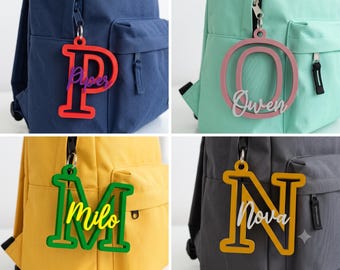May include: Four backpack charms featuring large, colorful, die-cut letters P, O, M, and N. Each letter is layered with a smaller, cursive-script name tag reading Piper, Owen, Milo, or Nova in contrasting colors, attached by a silver metal ring.