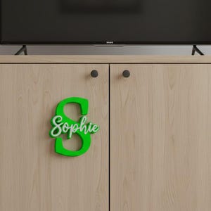 May include: A green wooden letter "S" with the name "Sophie" in white script attached to a light wood cabinet door. The cabinet has black knobs and is part of a media console. A television is above the cabinet.