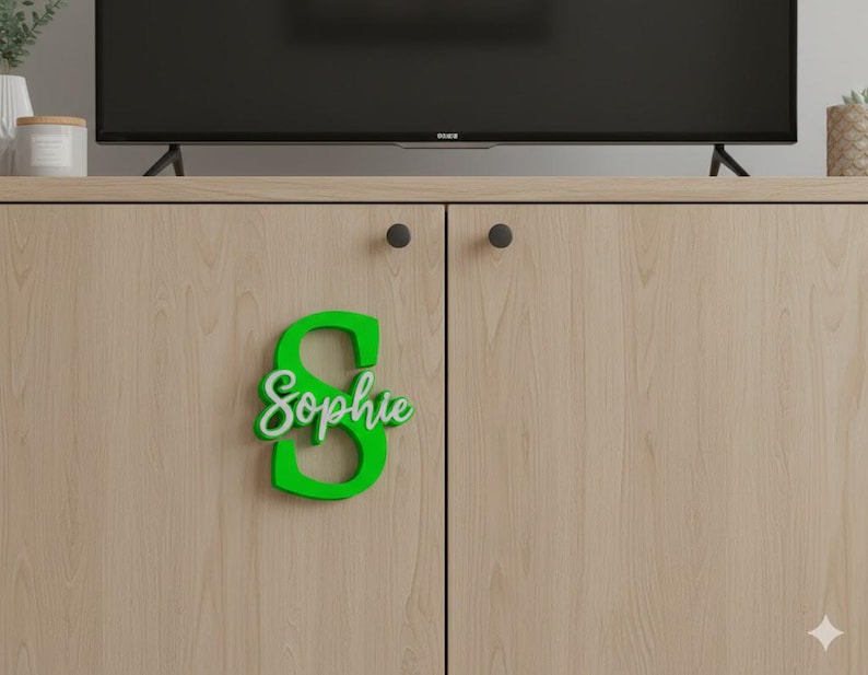 May include: A green wooden letter "S" with the name "Sophie" in white script attached to a light wood cabinet door. The cabinet has black knobs and is part of a media console. A television is above the cabinet.