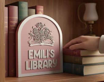 Puede incluir: Un letrero de madera rosa y blanco que dice "EMILI'S LIBRARY" con un diseño floral. El letrero está en un estante de madera con libros. Una mano alcanza una pila de libros.