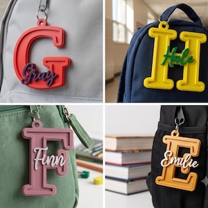 Pode incluir: Quatro etiquetas de mochila com as letras maiúsculas G, H, F e E nas cores vermelha, amarela, rosa e laranja. Cada etiqueta exibe um nome personalizado em letra cursiva: Gray, Hale, Finn e Emilie, presas em diferentes mochilas.